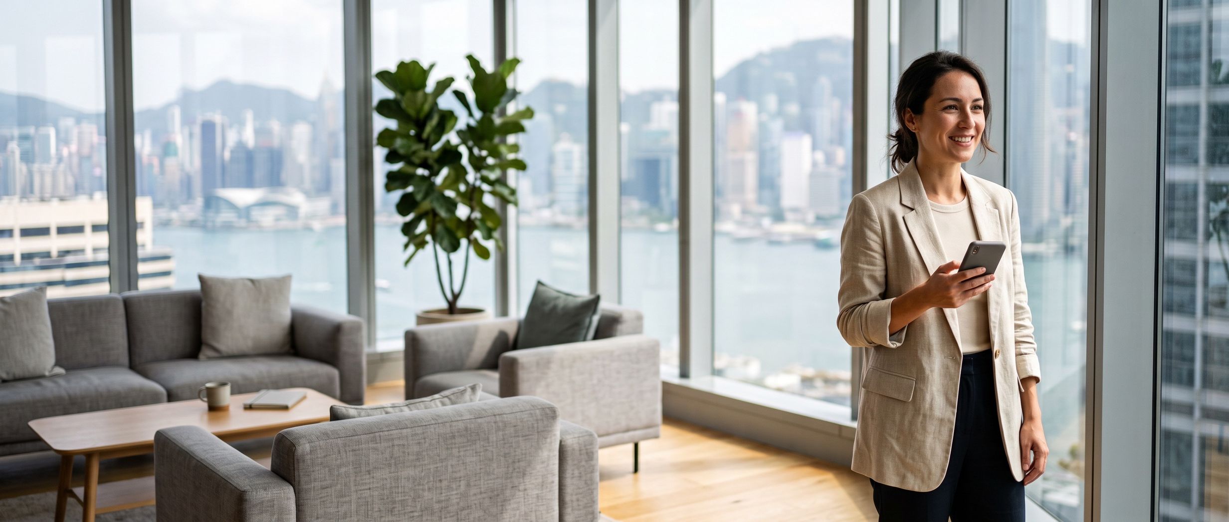 A woman in a Hong Kong office lounge, phone in hand, the harbour through the glass behind her
