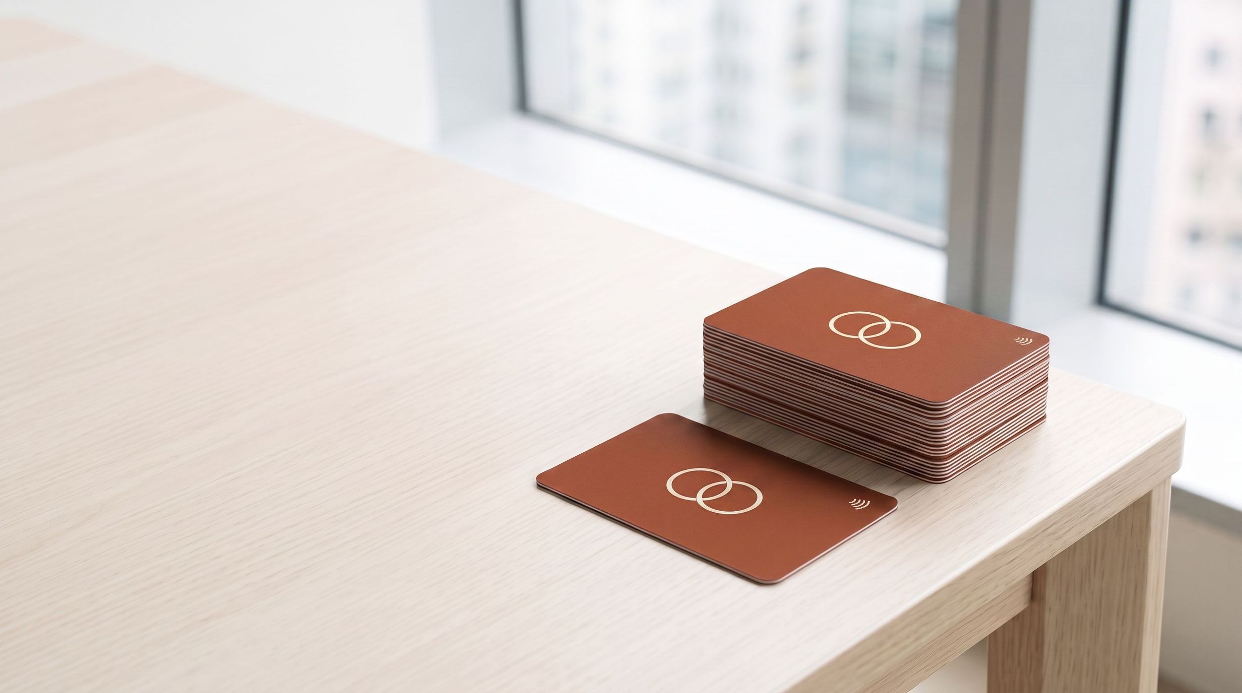 A squared stack of terracotta company cards printed with a cream two-circle logo, on a pale oak table by the window, one card lying face-up in front